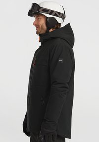 Zwarte skijack met een capuchon, voorzien van oranje accenten, ritszakken en een logo-opdruk op de mouw. Ontworpen voor wintersport.