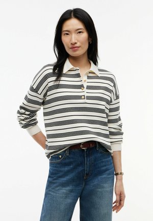 Kvinde med langt sort hår iført en hvid og sort stribet langærmet polo skjorte og blå jeans, stående med den ene hånd på hoften.