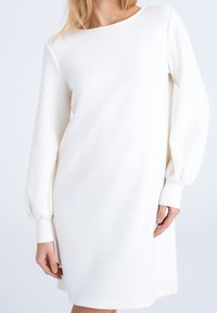 Vit sweatshirtklänning med långa puffärmar och rundad halsringning. Tyget verkar mjukt med en jämn yta och en lätt lyster.