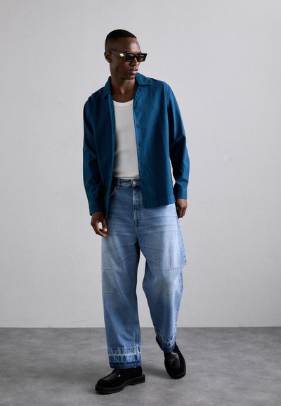 Chemise boutonnée teal par-dessus un débardeur blanc, associée à un jean palazzo bleu clair. Présente des couleurs unies et une coupe décontractée. Chaussures à plateforme noires.