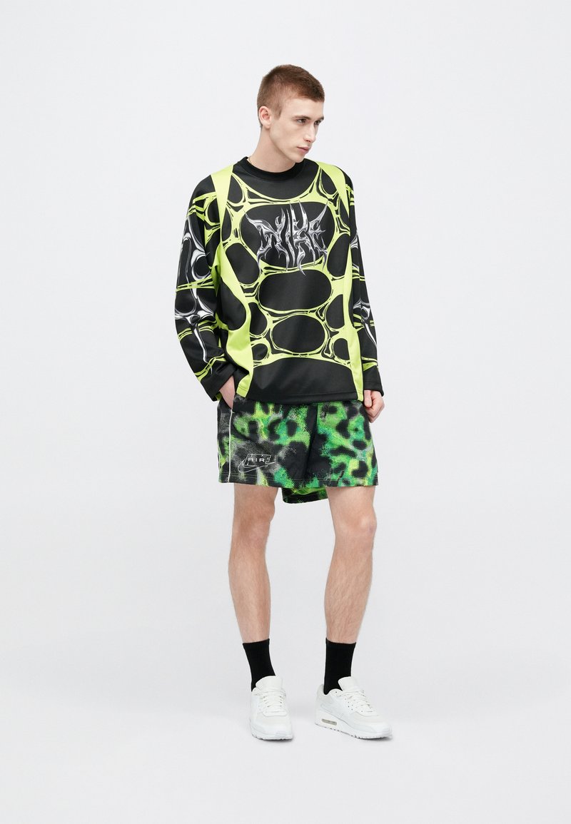 Oversized black sweatshirt with neon green designs, paired with green patterned shorts. White sneakers and black socks complete the outfit.