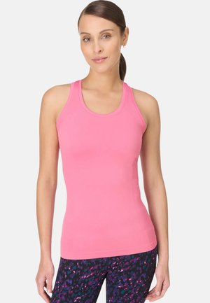 Vrouw met bruin haar in een paardenstaart, gekleed in een mouwloos roze tanktop en donkere, met patronen bedrukte leggings, staand tegen een effen lichte achtergrond.