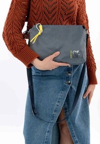 Borsa a tracolla grigia con tessuto texturizzato, dettagli gialli, accessori in argento e tracolla regolabile, appoggiata su una gonna di jeans.