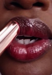 Läppstift i en metallisk roséguldig tub som appliceras på djup röda, glansiga läppar. Tänder synliga, slät yta och rik färgkontrast mot huden.