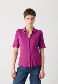 Tett, kortermet skjorte i lys rosa stoff med knappestolpe, klassisk krage og glatt tekstur. Kombinert med blå jeans.
