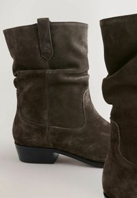 Bottines en suède marron à design flou, bout rond et petit talon noir. Présente des coutures discrètes et des coutures latérales pour le détail.