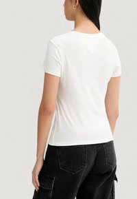 Bílé přiléhavé tričko s krátkými rukávy a kulatým výstřihem, zobrazené zezadu, doplněné černými cargo kalhotami s kapsami.