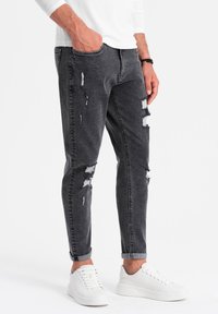 Hombre que viste jeans negros deslavados y rasgados, con los tobillos enrollados, y zapatillas blancas, de pie con una mano en el bolsillo frente a un fondo sencillo.