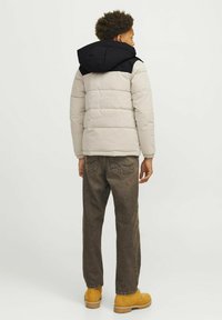 Pufota jaka ar beiga ķermeni un melnu kapuci, ar quilted tekstūru. Apvienota ar brūniem džinsiem un dzeltenām zābakiem, skatīta no aizmugures.