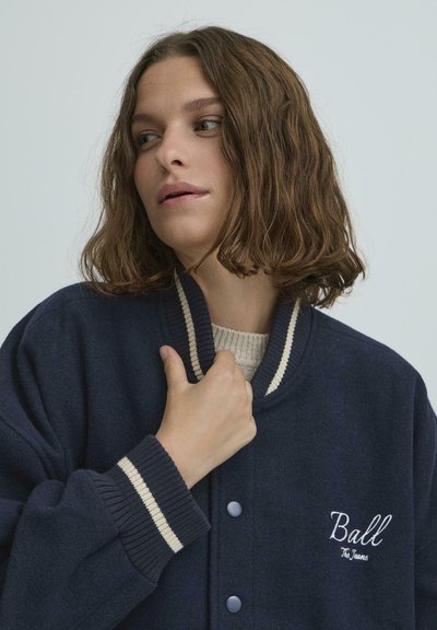 Jachetă bleumarin dintr-un amestec de lână, cu guler și manșete cu nervuri, decorate cu dungi crem. Include text brodat "Ball" pe piept.