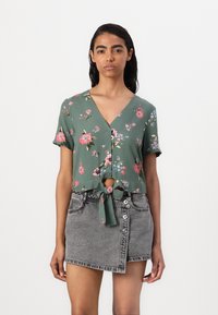 Top corto floreale verde con maniche corte, scollatura a V profonda e dettaglio con fiocco in vita, abbinato a una mini gonna in denim grigia con chiusura a bottoni.