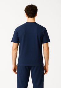 Mężczyzna o kręconych włosach, ubrany w jednolitą granatową koszulkę z krótkim rękawem i pasujące spodnie, stoi tyłem na białym tle.