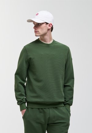 Młody mężczyzna w zielonej bluzie i spodniach do kompletu, białej czapce z daszkiem z czerwonym logo, stoi z rękami w kieszeniach, patrząc w bok.