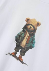 Camiseta blanca con un gráfico de un oso con capucha marrón y chaqueta verde azulado, con zapatos naranjas, de pie con las manos en los bolsillos.