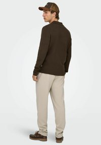 Brązowy sweter z kołnierzykiem, beżowe spodnie dresowe oraz brązowe buty. Model nosi brązową czapkę z siateczką z tyłu, z logo z przodu.