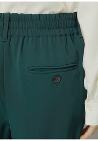 Grüne Hose aus strukturierter Baumwolle mit elastischem Bund und einer kleinen Seitentasche mit einem schwarzen Knopfdetail.