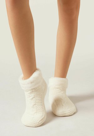 Jambes portant des chaussettes de cheville tricotées blanches avec un poignet doux et duveteux sur un fond clair.