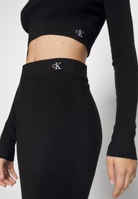 Černý žebrovaný set skládající se z crop topu a sukně v upnutém designu, s logem na elastických pasech a manžetách rukávů.