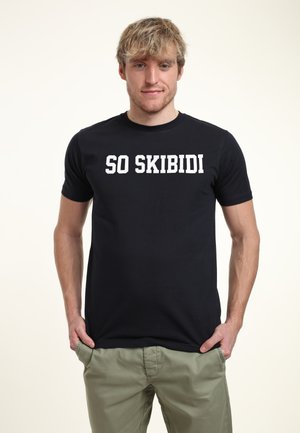 Sort bomuld t-shirt med teksten "SO SKIBIDI" i fede hvide bogstaver. Kortærmet og med rund halsudskæring. Båret med lysegrønne bukser.