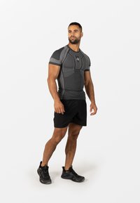 Atletski moški stoji, oblečen v oprijeto črno-sivo športno majico, črne kratke hlače in črne tekaške copate, na belem ozadju.