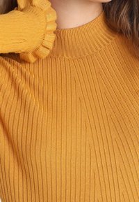 Raitapintainen sinapin keltainen villapaita, jossa on korkea kaulus ja röyhelöhihoja, ominaispiirteenä tekstuurinen neulontakuvio kankaassa.