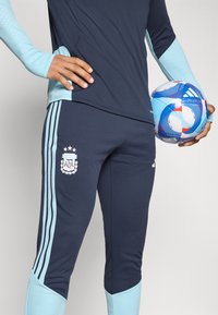 Άτομο με ναυτικές και ανοιχτό μπλε αθλητικές ενδυμασίες κρατάει μια μπλε και λευκή ποδοσφαιρική μπάλα της Adidas στο ισχίο του.