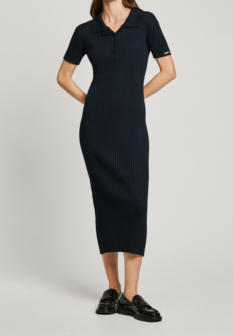 Femme portant une robe midi en maille côtelée noire à manches courtes, avec un col boutonné, et des mocassins noirs, debout devant un fond uni.