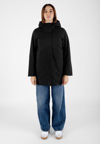 Parka nera impermeabile con cappuccio, caratterizzata da un taglio dritto e chiusura a pressione. Abbinata a jeans in denim blu a gamba larga e scarpe da ginnastica bianche.
