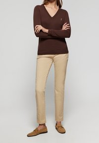 Pull en maille marron à col en V, pantalon beige ajusté et mocassins marron. Design simple avec un détail subtil de logo sur le pull.
