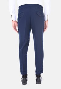 Pantaloni blu navy su misura con pieghe, due tasche posteriori con bottoni e orli con risvoltino. Indossati con scarpe nere e calze nere.