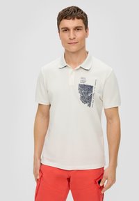 Weißes Polohemd aus Baumwolle mit einem navyblauen Grafikdesign auf der linken Brust und einem klassischen Kragen. Getragen mit korallenfarbenen Cargo-Shorts.