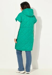 Quiltad grön väst med huva, korta ärmar och dragkedja. Har ett diamantmönster och bärs över en vit långärmad topp och blå jeans.