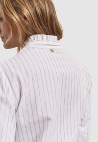 Biała koszula z cienkimi złotymi paskami, falbankowym kołnierzykiem oraz zapięciem na guziki z tyłu. Gładka tekstura, długie rękawy i luźny krój.