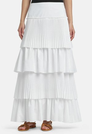 Kobieta w długiej, białej, warstwowej spódnicy z plisami i falbanami, w brązowych, płaskich sandałach na paski, stoi na tle gładkiego tła.