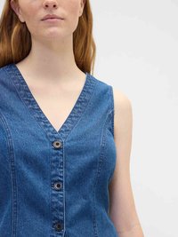 Woman with light skin and long red hair wearing a sleeveless blue denim button-up top against a plain light background.