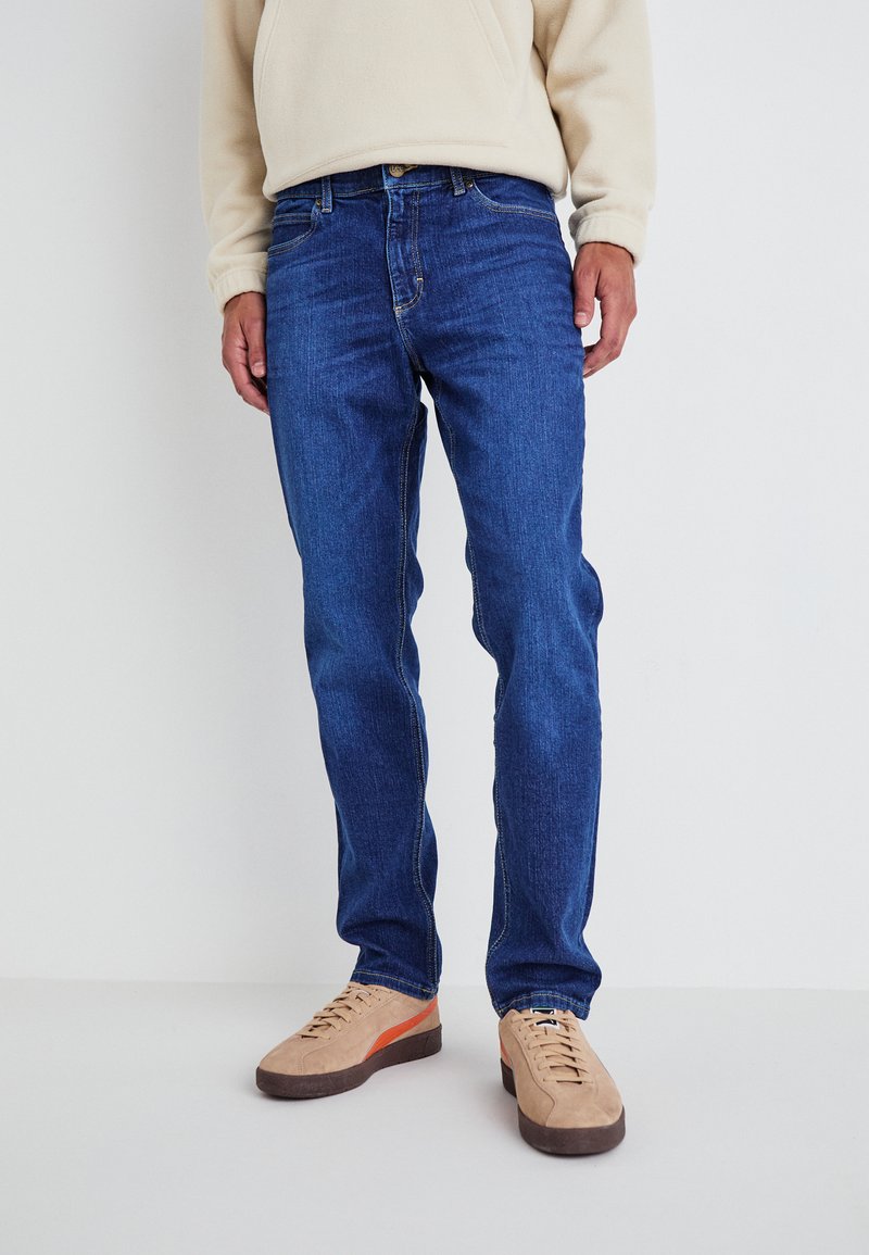 Vaqueros de mezclilla azul con un corte recto, que cuentan con cinco bolsillos, costuras amarillas y una textura suave. Llevados con zapatillas beige.