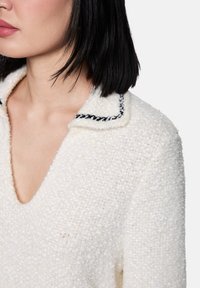 Frau mit schwarzen Haaren, die einen strukturierten weißen Pullover mit weitem V-Ausschnitt und einem Kragen trägt, der mit schwarzer Naht verziert ist.