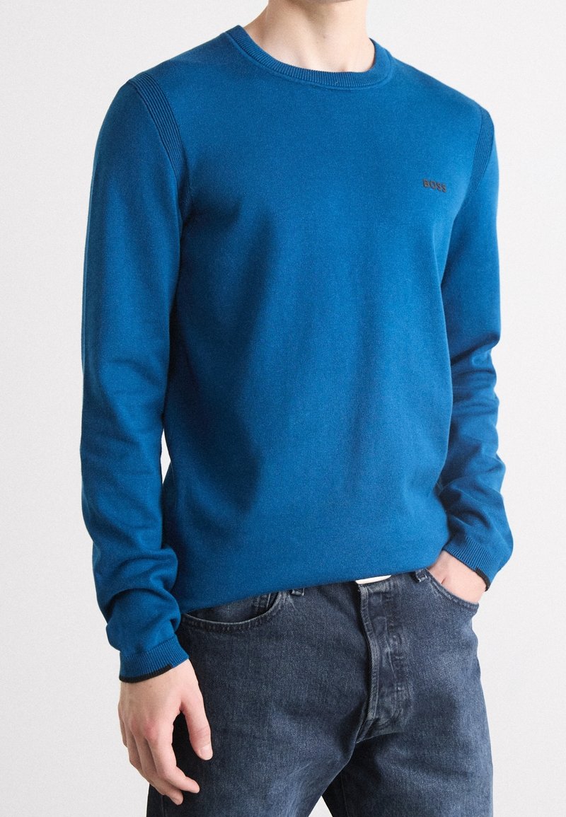 Homme portant un pull bleu ajusté à manches longues avec des poignets côtelés et un jean foncé, une main dans la poche.