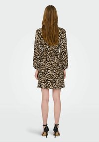 Frau mit langen Haaren, die ein knielanges Kleid mit Leopardenmuster und schwarze High Heels trägt, steht mit dem Rücken zu einem einfarbigen Hintergrund.