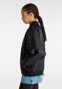 Zwarte bomberjack met een geerte zak op de mouw, ribbelmanchetten en zoom; gladde textuur; gelaagd over een blauw shirt.