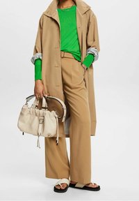 Kreemi trench coat päällä vihreän pitkähihaisen paidan, vaaleat leveälahkeiset housut, mustat sandaalit ja kreemi käsilaukku hihnoilla.