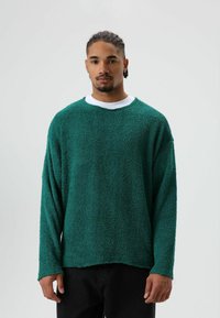 Groene gebreide trui met een zachte, pluizige structuur. Heeft een ronde halslijn en lange mouwen, gedragen over een wit topje. Donkere broek zichtbaar.