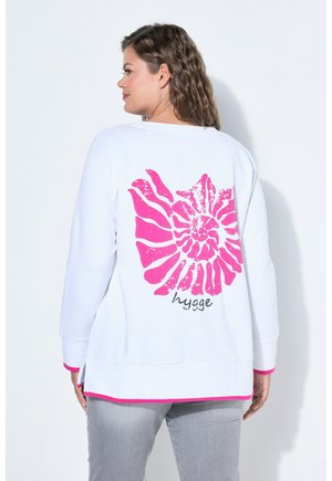 Vrouw met golvend haar draagt een wit top met lange mouwen, met een groot roze spiraalschelpontwerp en het woord "hygge" op de achterkant.