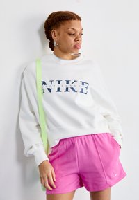 Bijela dukserica s plavim "NIKE" logotipom, nošena preko ružičastih kratkih hlača. Ima okrugli izrez i opušteni kroj. Zeleni remen preko ramena.