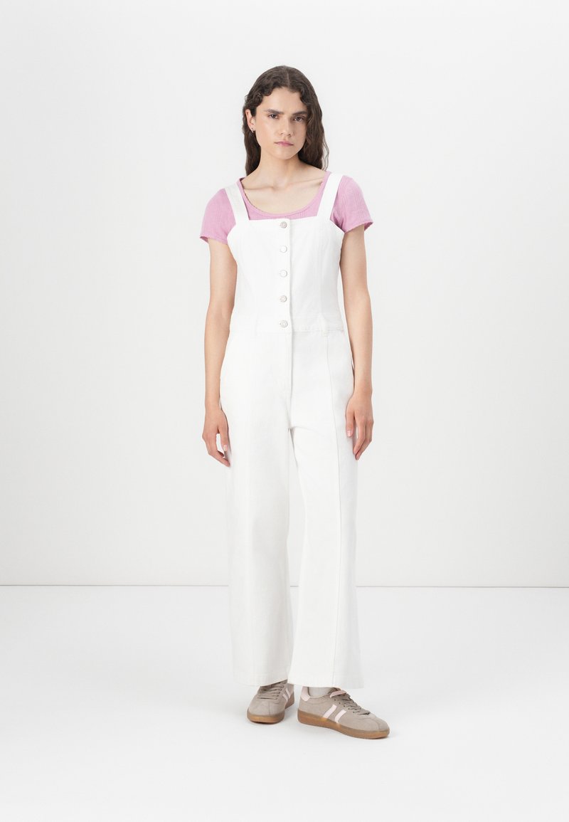 Overoles blancos de piernas anchas con un corsé ajustado y cuatro botones al frente, combinados con una blusa de manga corta rosa claro. Zapatillas color caramelo completan el look.