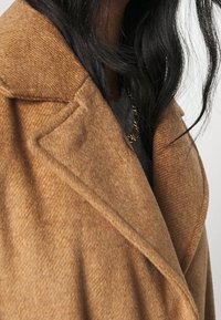 Cappotto in misto lana marrone con colletto a risvolto, tessuto testurizzato e tasche laterali inclinate, che copre parzialmente una maglia grigia sottostante.