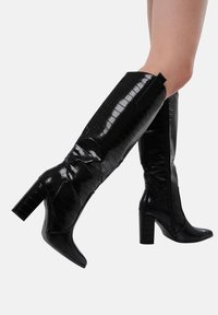Botas negras hasta la rodilla en imitación de cuero con relieve de cocodrilo. Presentan un tacón grueso y una cremallera lateral para el cierre, con un acabado brillante y elegante.