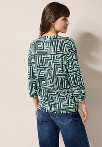 Mintgrünes Oberteil mit schwarzem geometrischen Muster, dreiviertelärmel und einer lockeren Passform, kombiniert mit dunkelblauen Jeans.