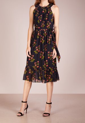 Femme portant une robe noire sans manches avec imprimé floral rose, ourlet volanté et ceinture nouée à la taille, associée à des talons noirs à bride à la cheville.