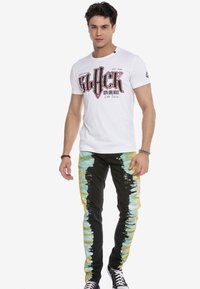 Weißes Baumwoll-T-Shirt mit auffälligem schwarzen und roten Grafikdruck, kombiniert mit schwarzen Denim-Jeans, die Batik-Akzente in Blau und Gelb aufweisen.